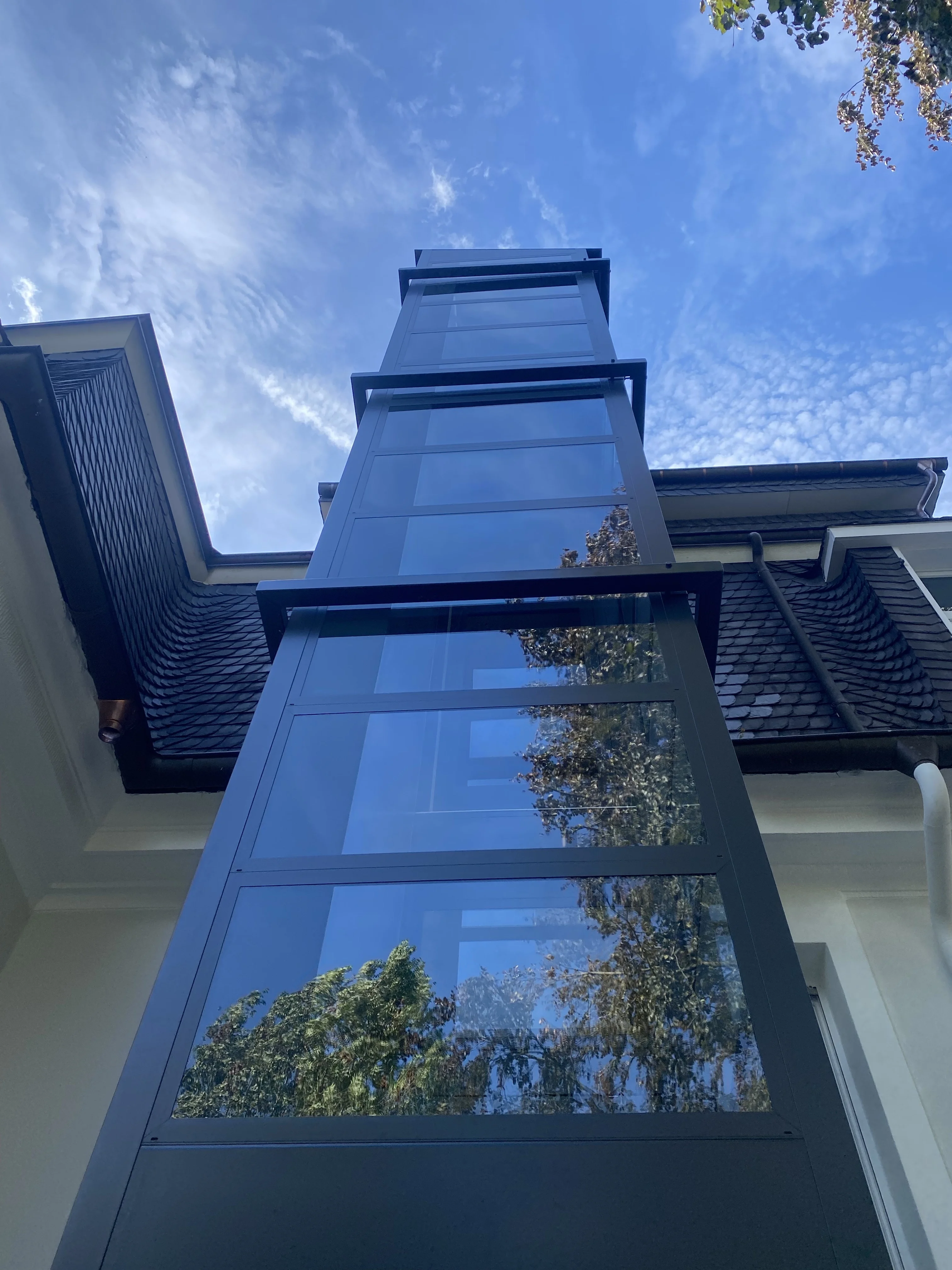Dunkler Glasschacht eines Außenaufzugs an einem Wohnhaus mit Schieferdach
